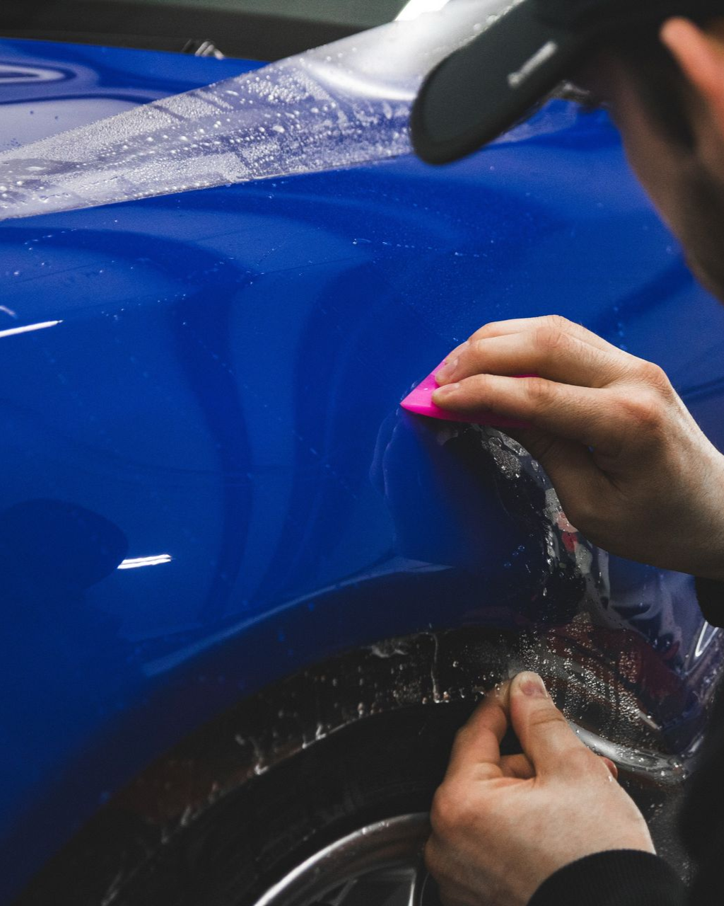 Hands apply protective film to a blue car panel with a squeegee.