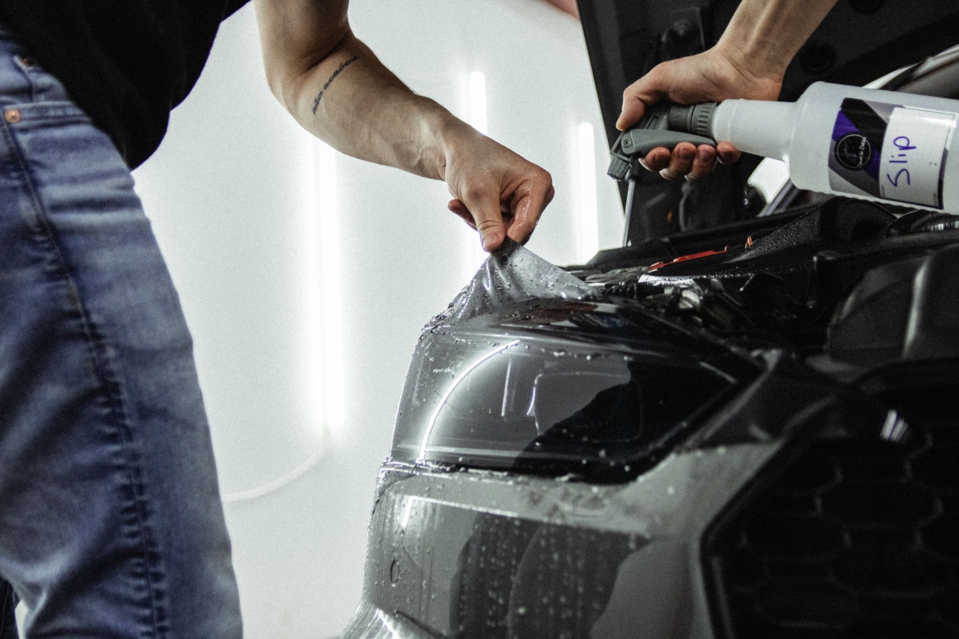 Hands applying protective film to the front of a gray car in a workshop