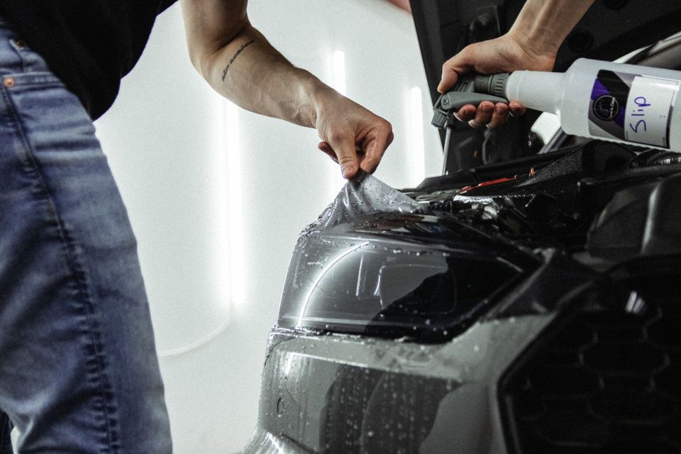 Two people applying protective film to a black car hood in a workshop