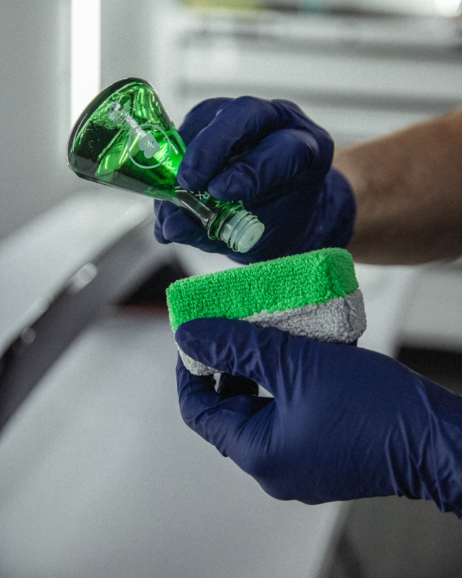 Gloved hands cleaning a green-and-gray sponge with a green spray bottle, close-up indoors