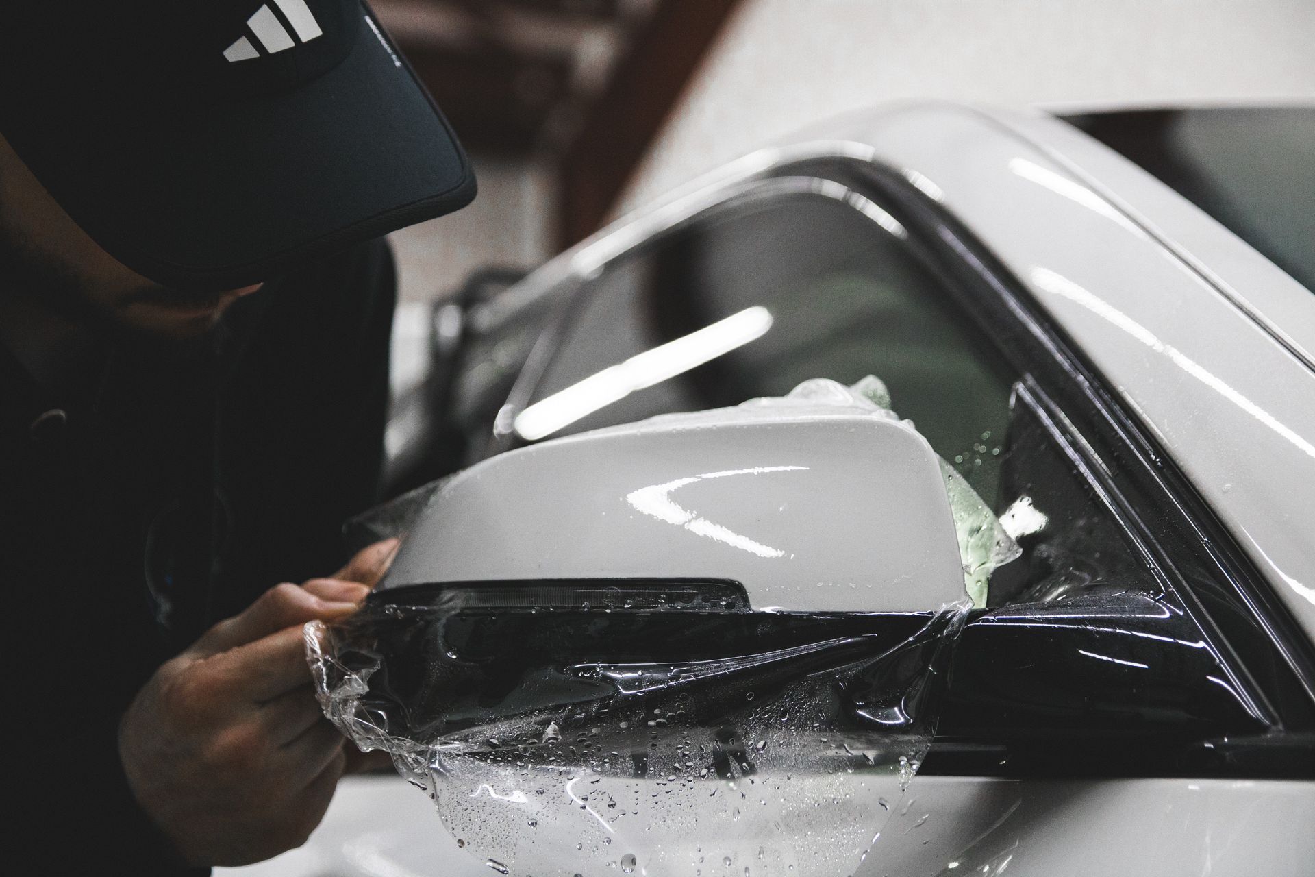 Person scrapes debris from a white car side mirror with a hand tool in a workshop