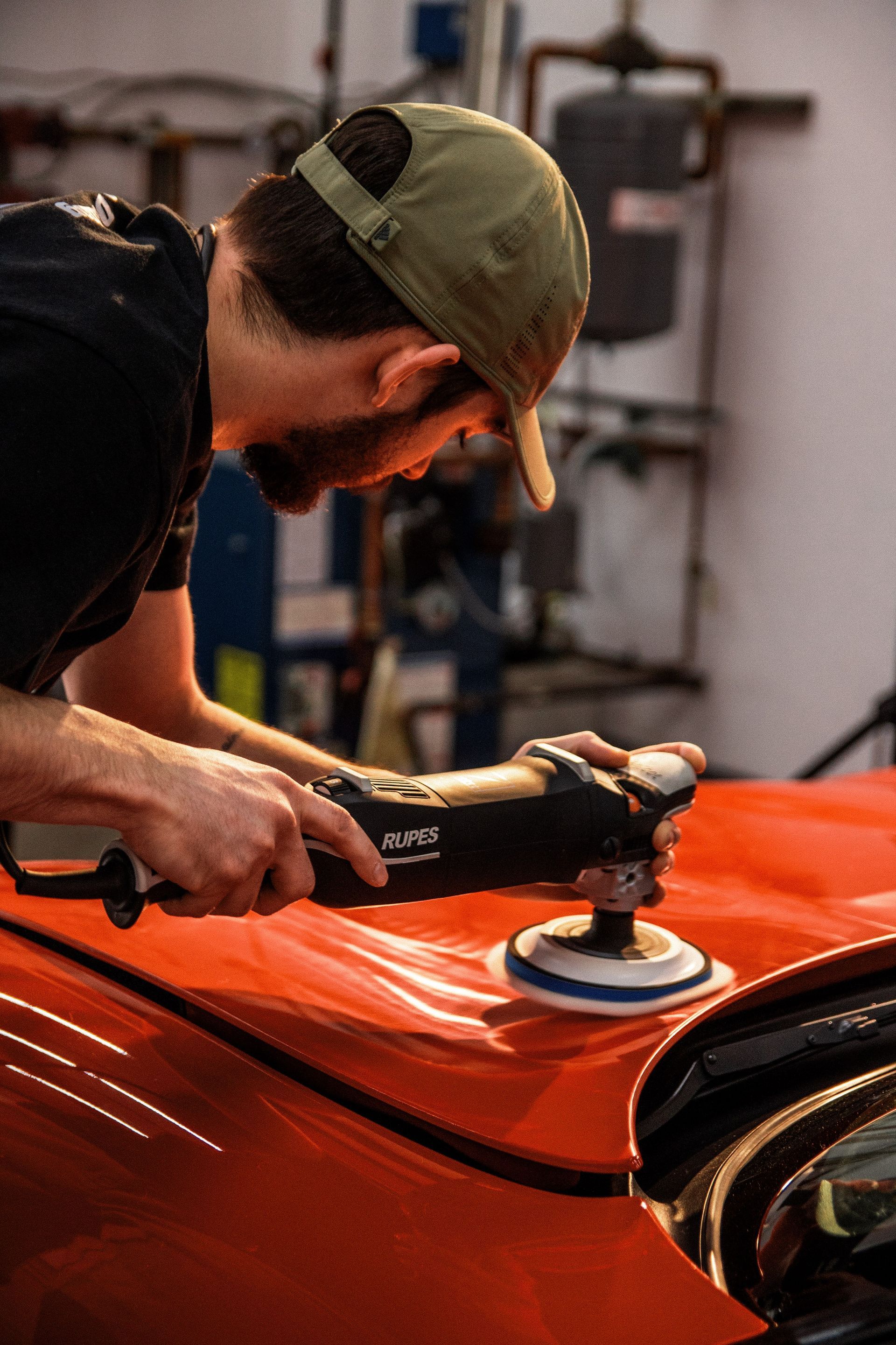 Person polishing a bright orange car hood with a buffer in a garage