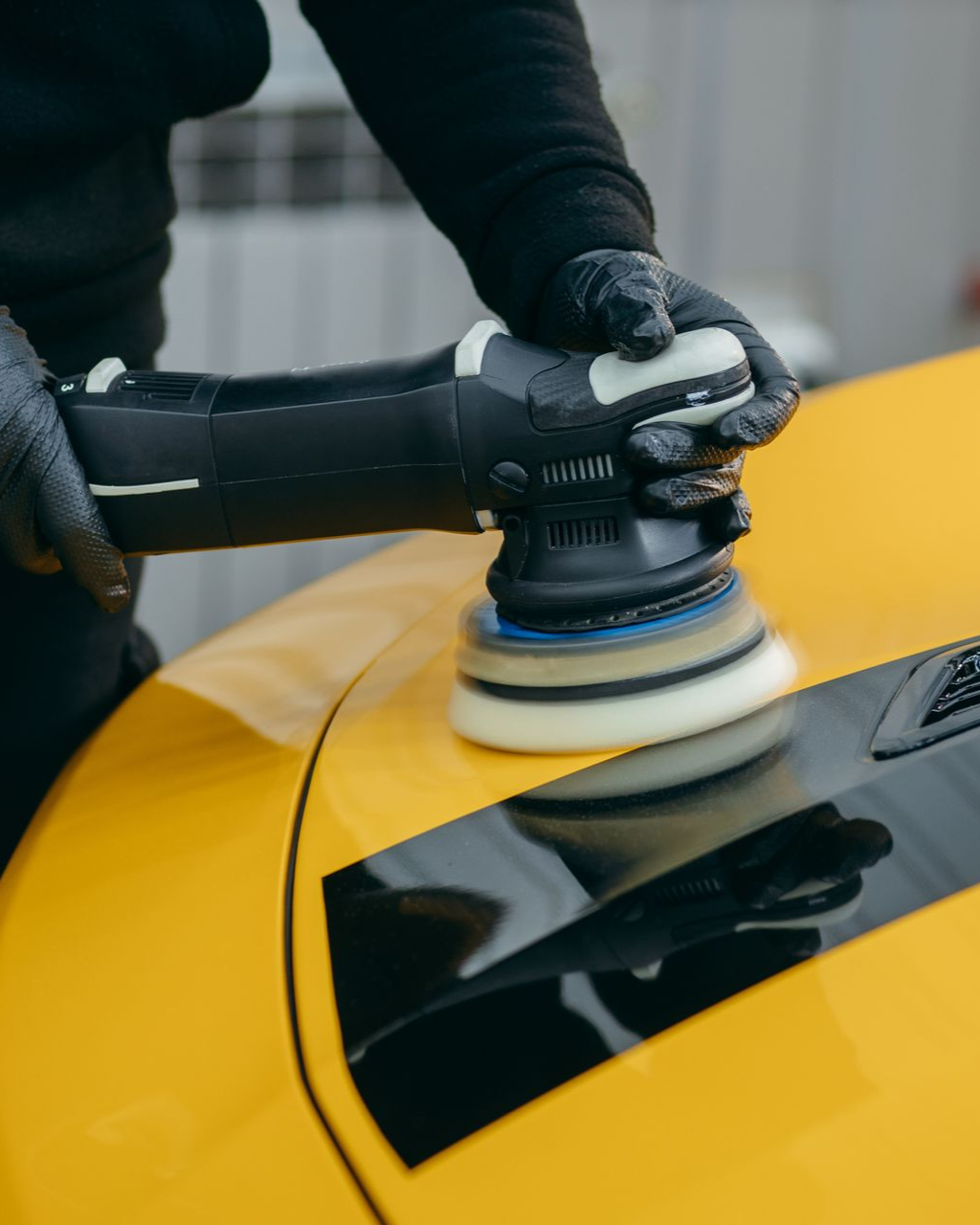 Person polishing a yellow car with a handheld buffer near a black window panel