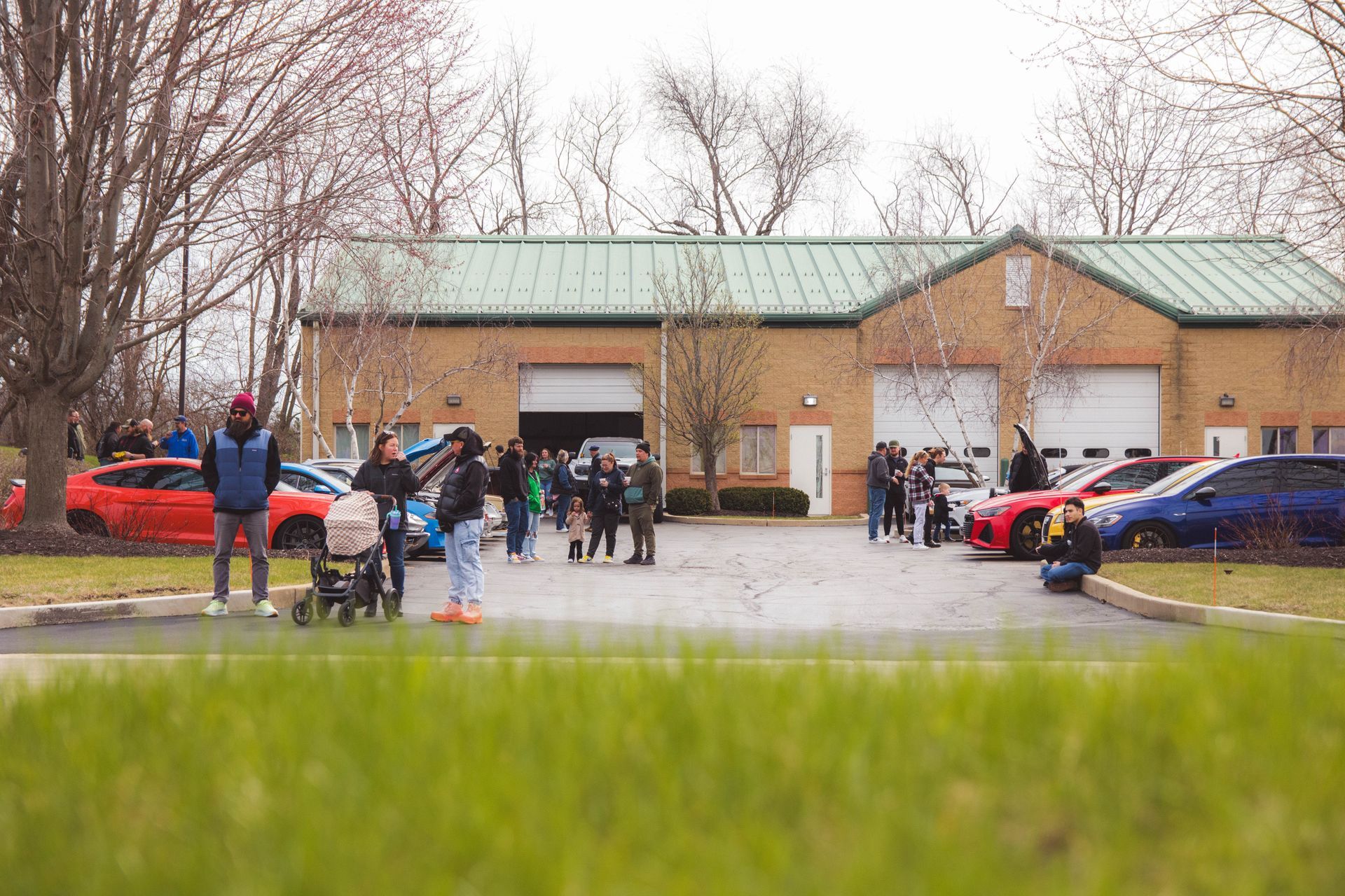 People gathered outside a brick building with a green roof, with cars parked nearby and grass in the foreground.