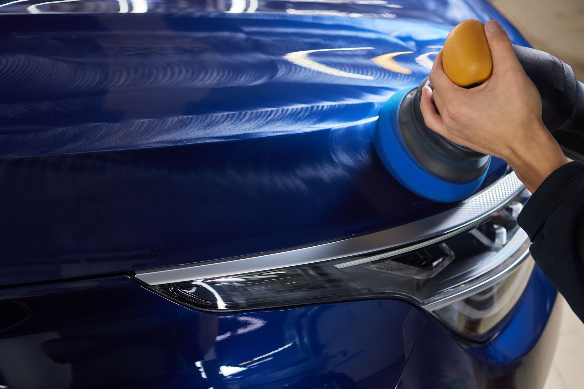 Hands polishing a blue car with a yellow buffer pad and cloth