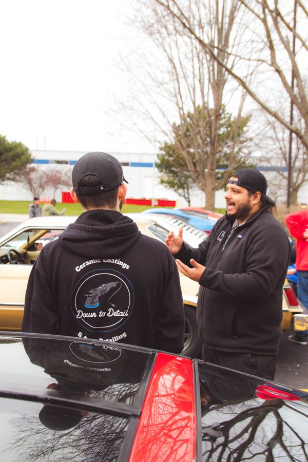 Two men talking beside a black car, one in a black hoodie with red accents, in a parking lot.