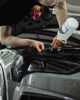 Person uses a spray bottle on a black car hood, wearing gloves in a garage.
