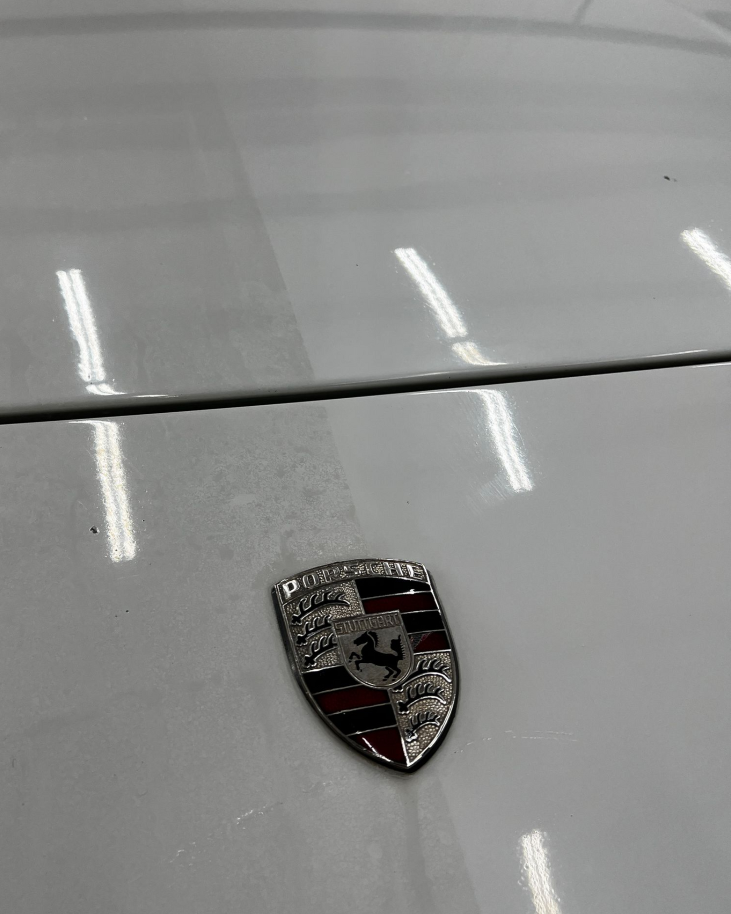 Porsche hood emblem on a silver car, with reflections of overhead lights.