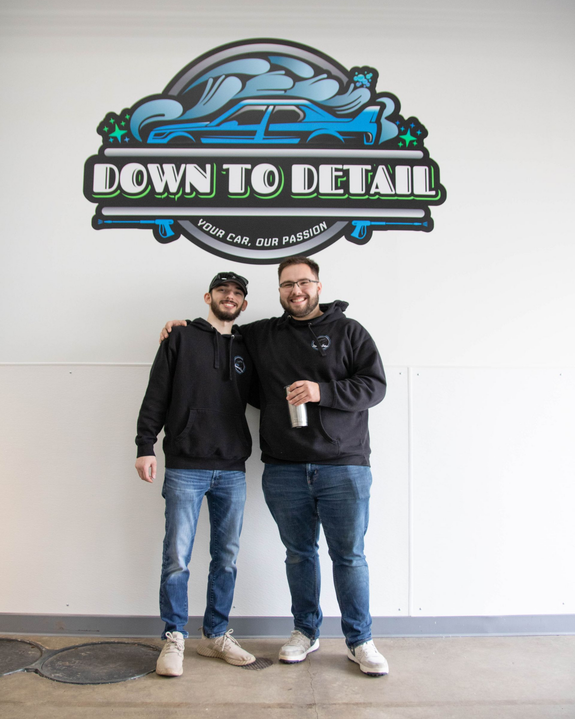 Two people standing under a “Down to Detail” sign, smiling and holding a drink in a white-walled room.