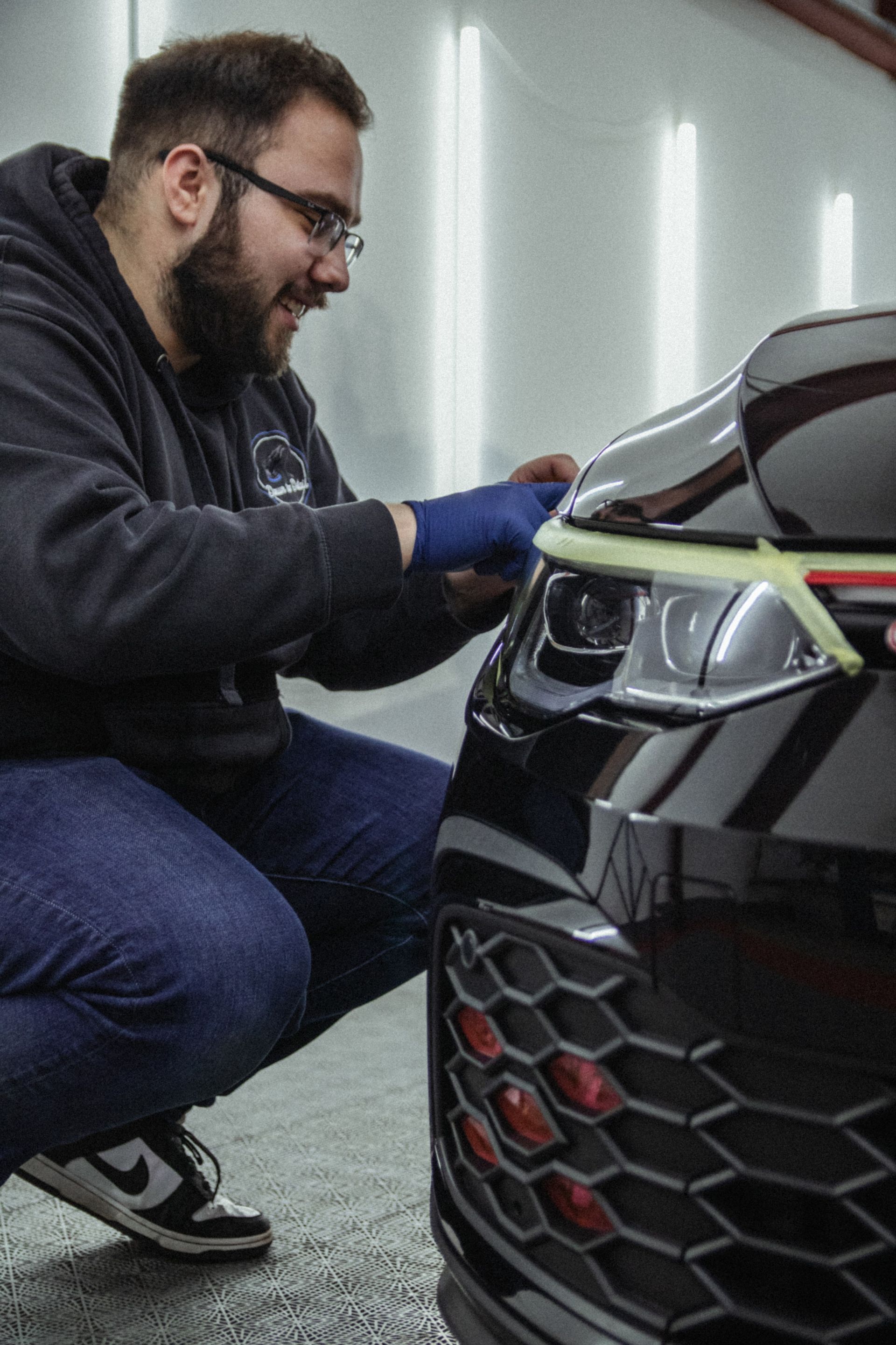Mechanic kneeling beside a car, inspecting the front headlight and grille in a garage