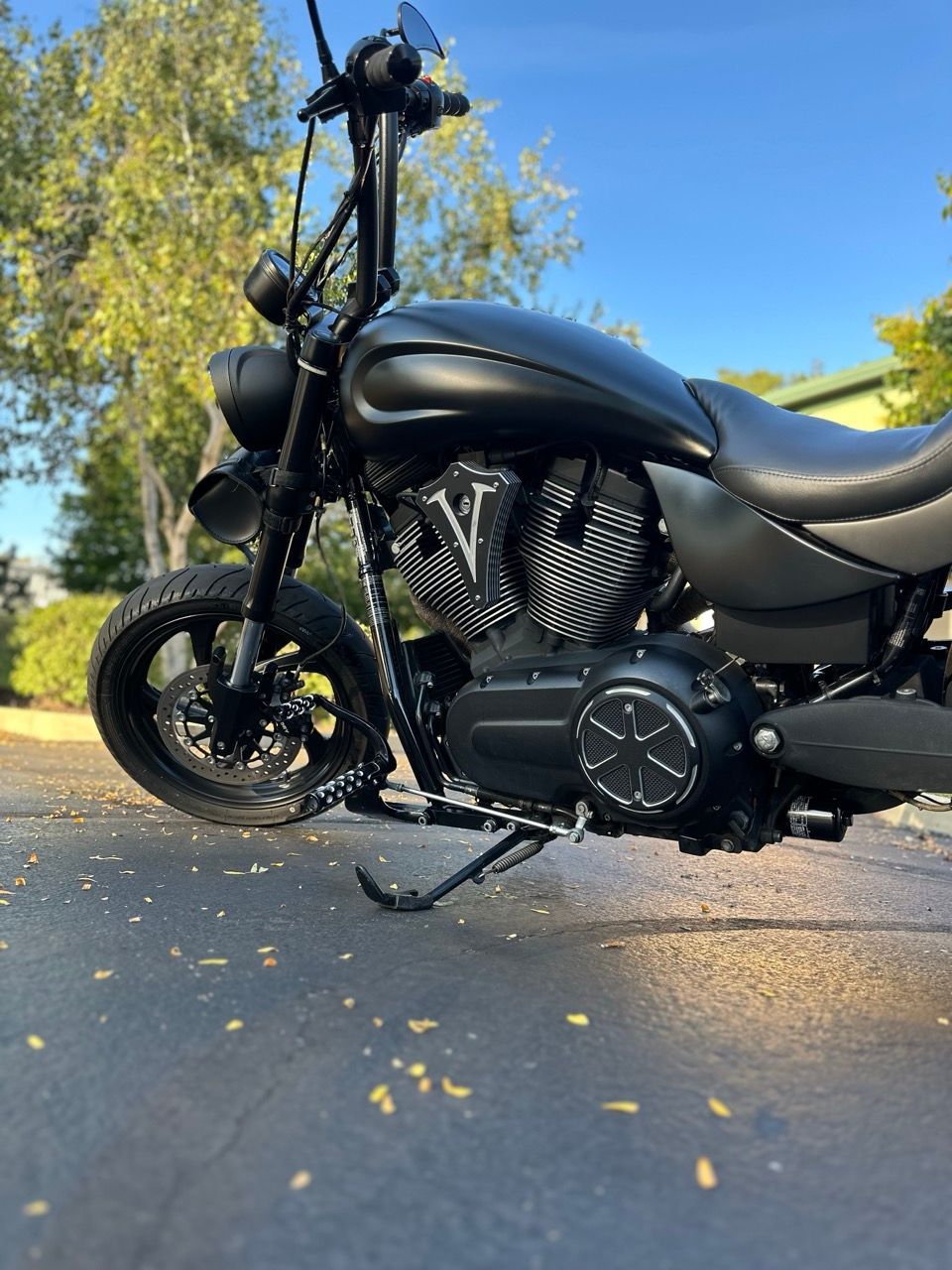 Black custom motorcycle parked on a leaf-strewn road under a blue sky with trees in the background