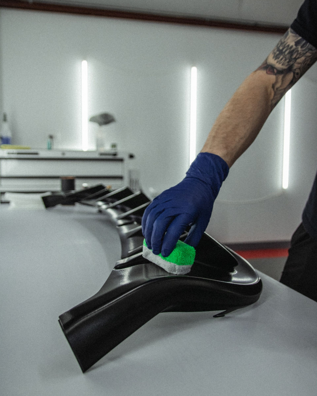 Gloved hand cleaning a black car part with a green sponge in a workshop