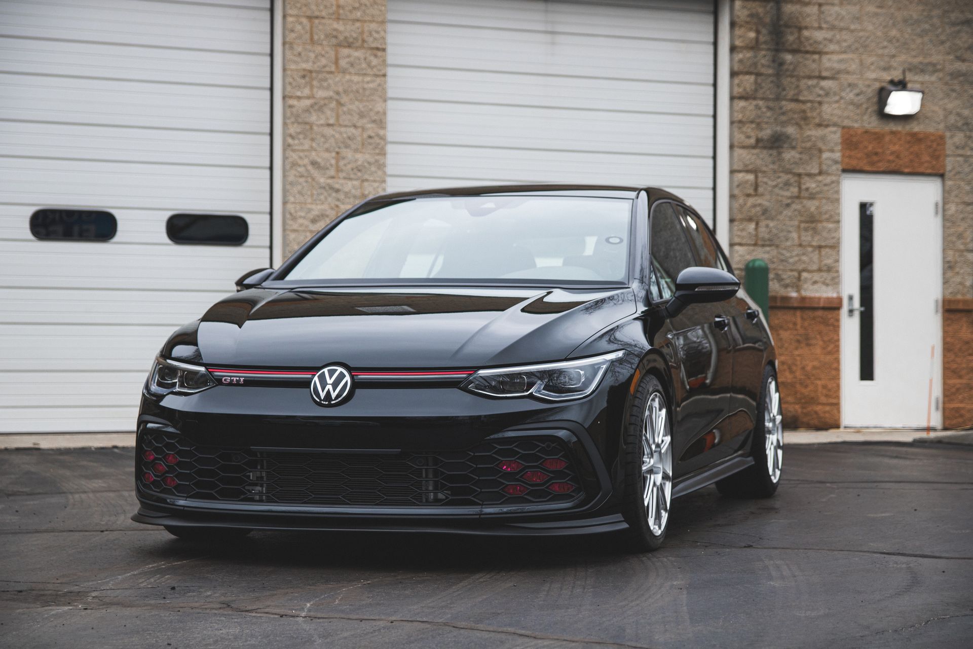 Black Volkswagen hatchback parked in front of a garage and brick building.