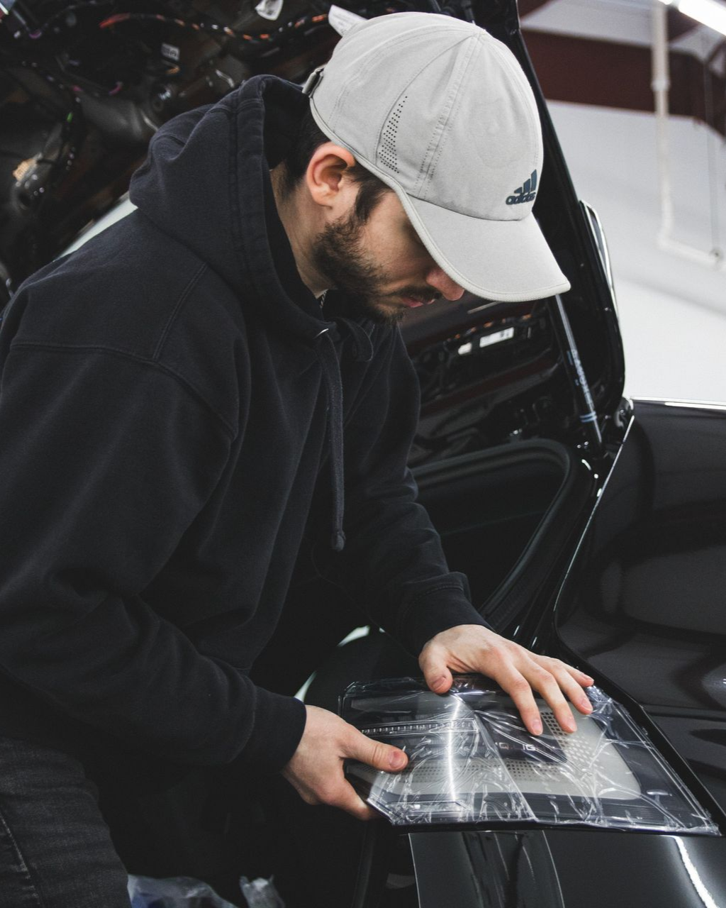 Person in a black hoodie and white cap inspects a reflective panel beside a car with the hood open