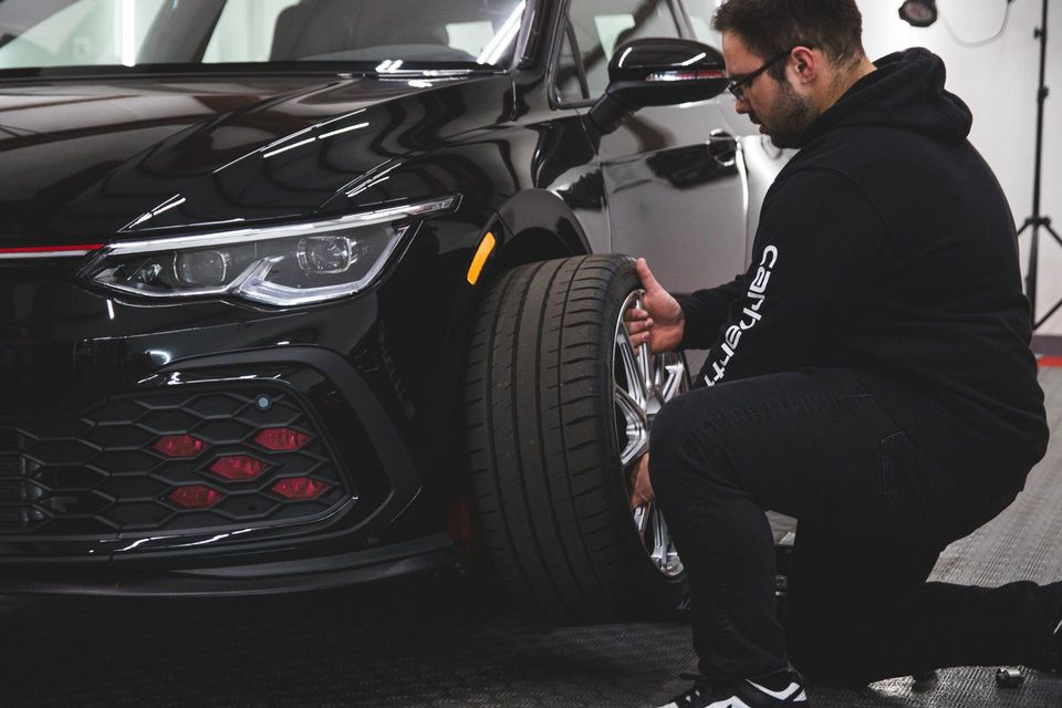Man inspecting and cleaning the front wheel of a black sports car in a garage