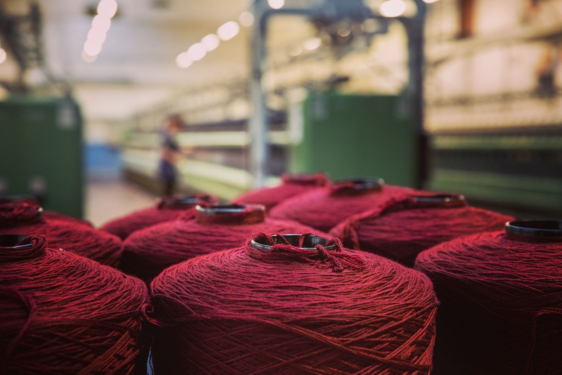In una fabbrica tessile, grandi bobine di filato rosso scuro sono allineate, con un operaio sfocato sullo sfondo.