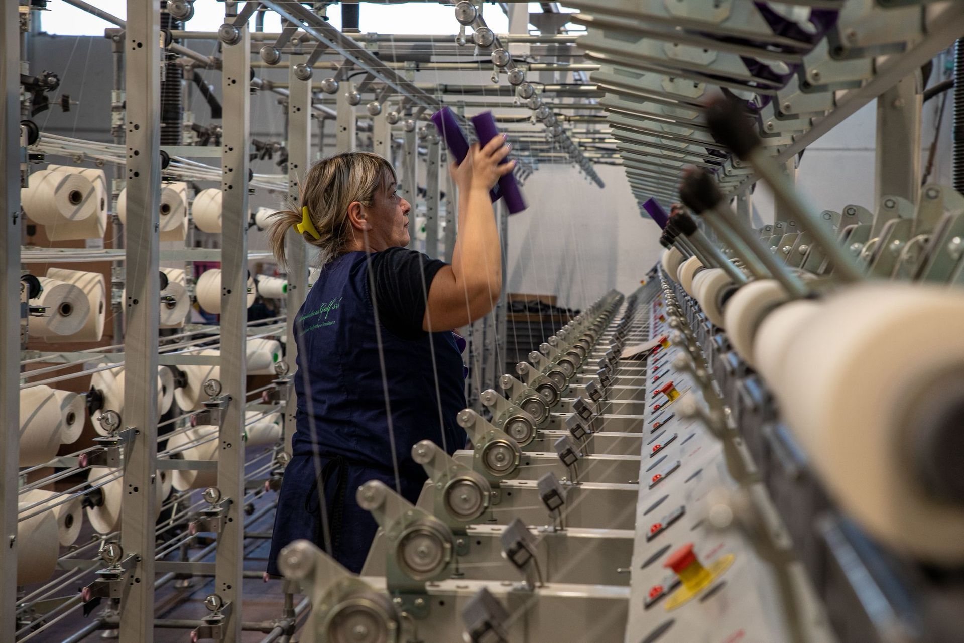 Un operaio in una fabbrica si occupa di una grande macchina tessile industriale piena di file di rocchetti di filato bianco.