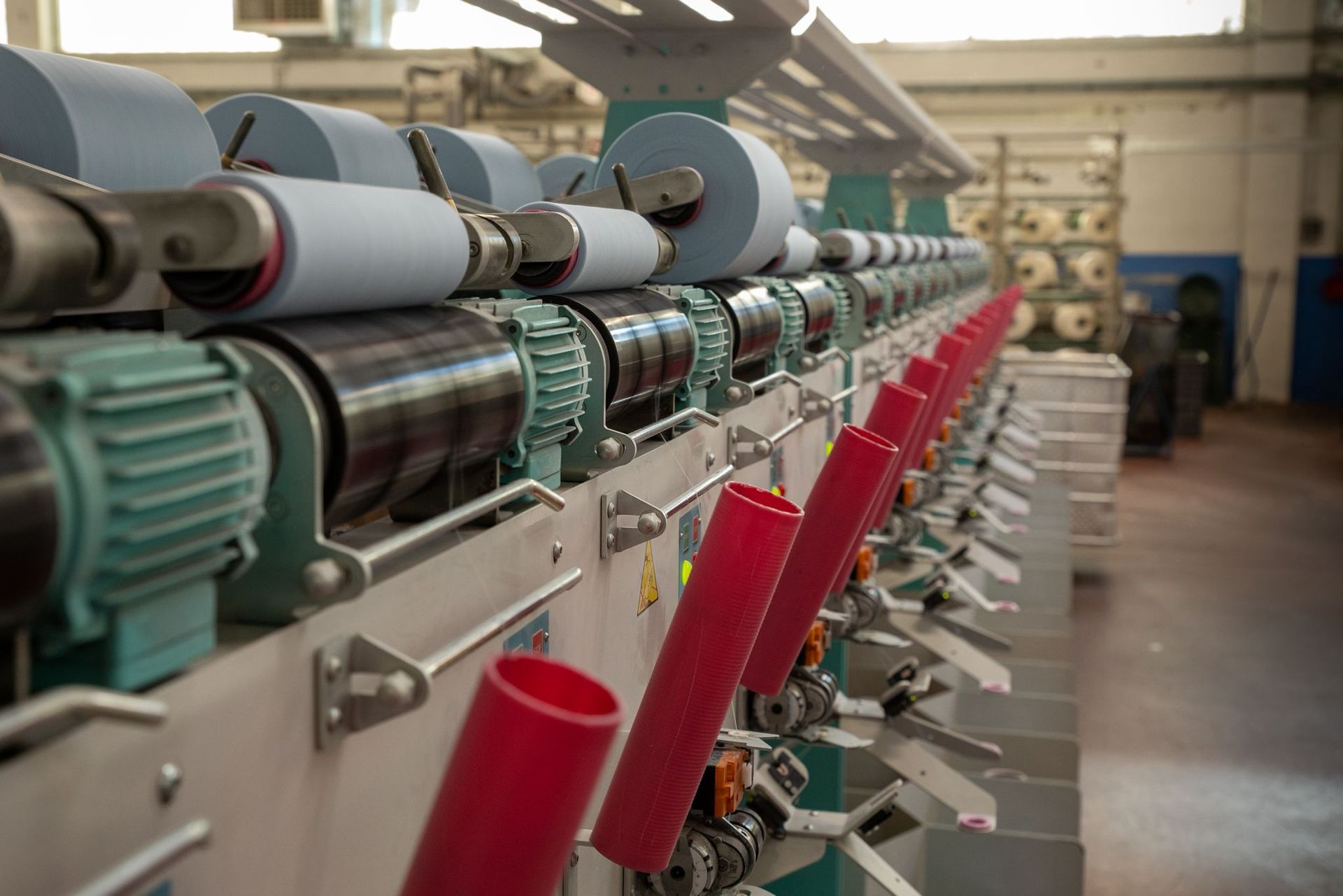 Macchina per filatura industriale in una fabbrica, con numerose bobine di filo azzurro e fusi di plastica rossa.
