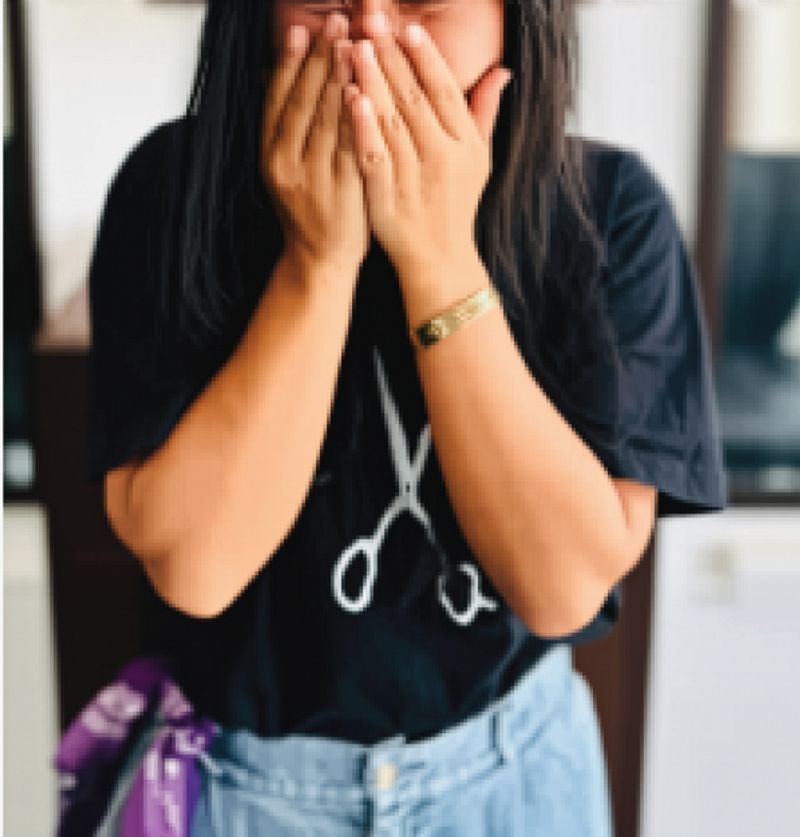 Woman with hands covering her mouth, wearing a black t-shirt with scissors, blue jeans, and a gold bracelet.