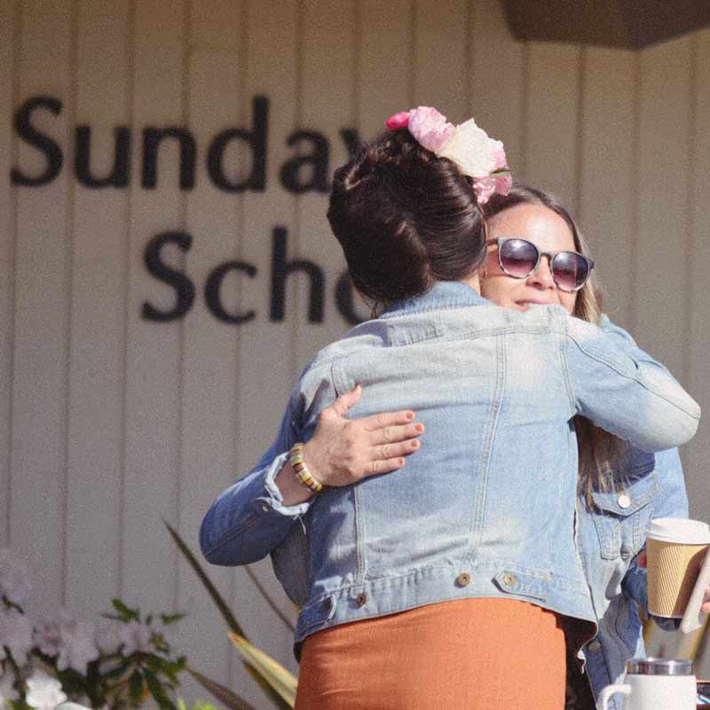 Two women hugging in front of a sign that says sunday school