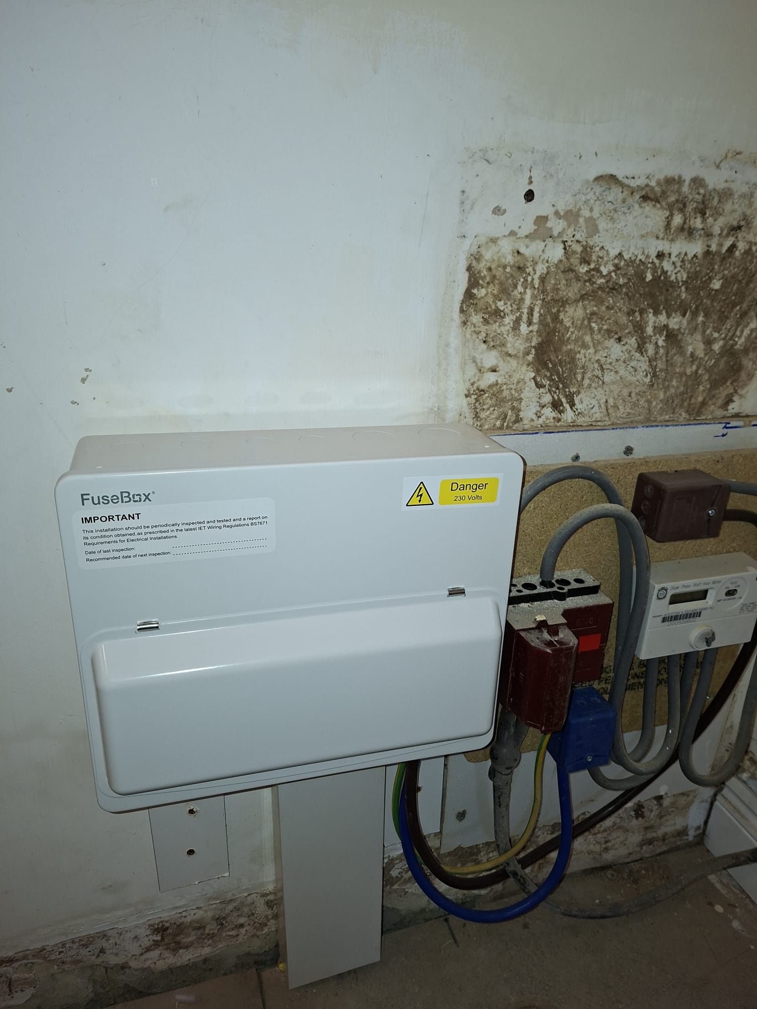 A white wall-mounted electrical consumer unit next to various electrical meters and cables on a rough, unfinished wall.