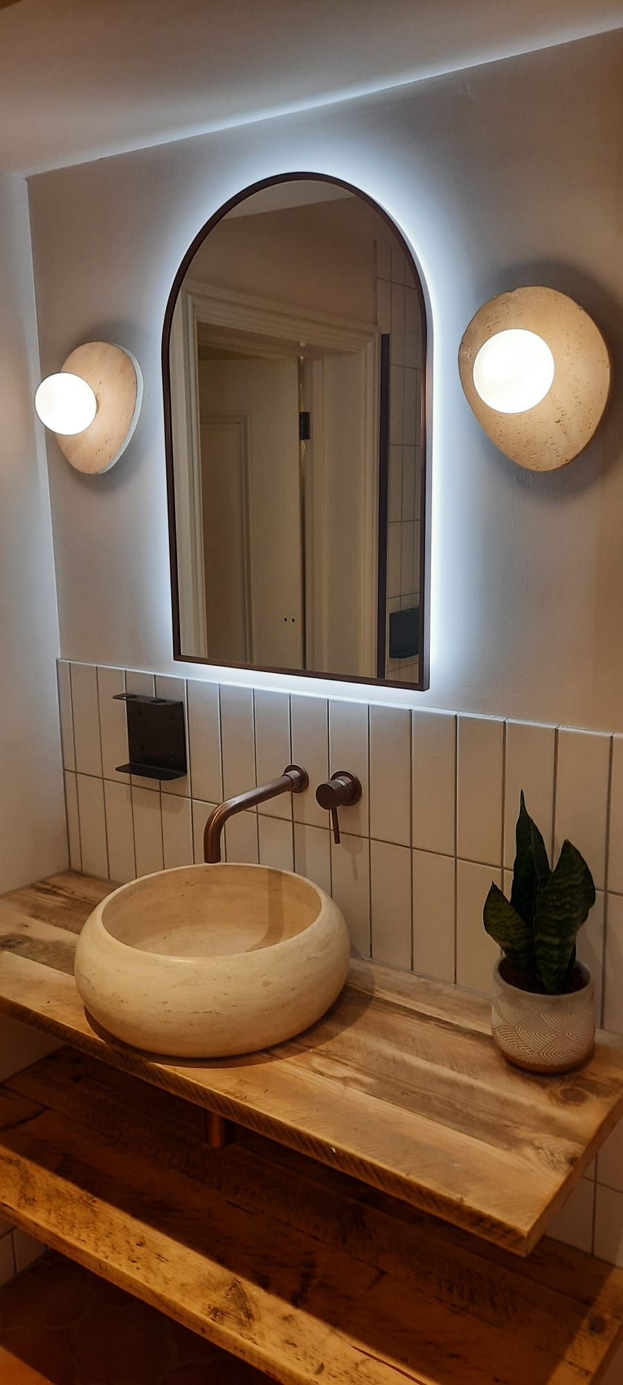 A modern bathroom vanity features a stone vessel sink, wooden countertop, arched backlit mirror, and two wall sconces.