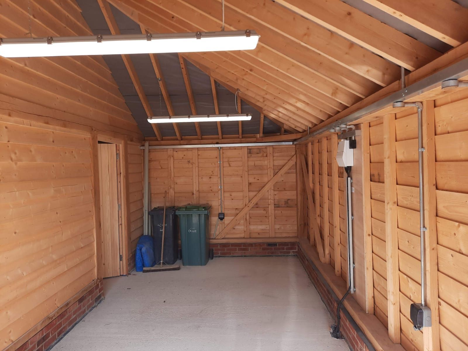 Interior of a wooden garage with two fluorescent lights, trash cans, and a concrete floor.