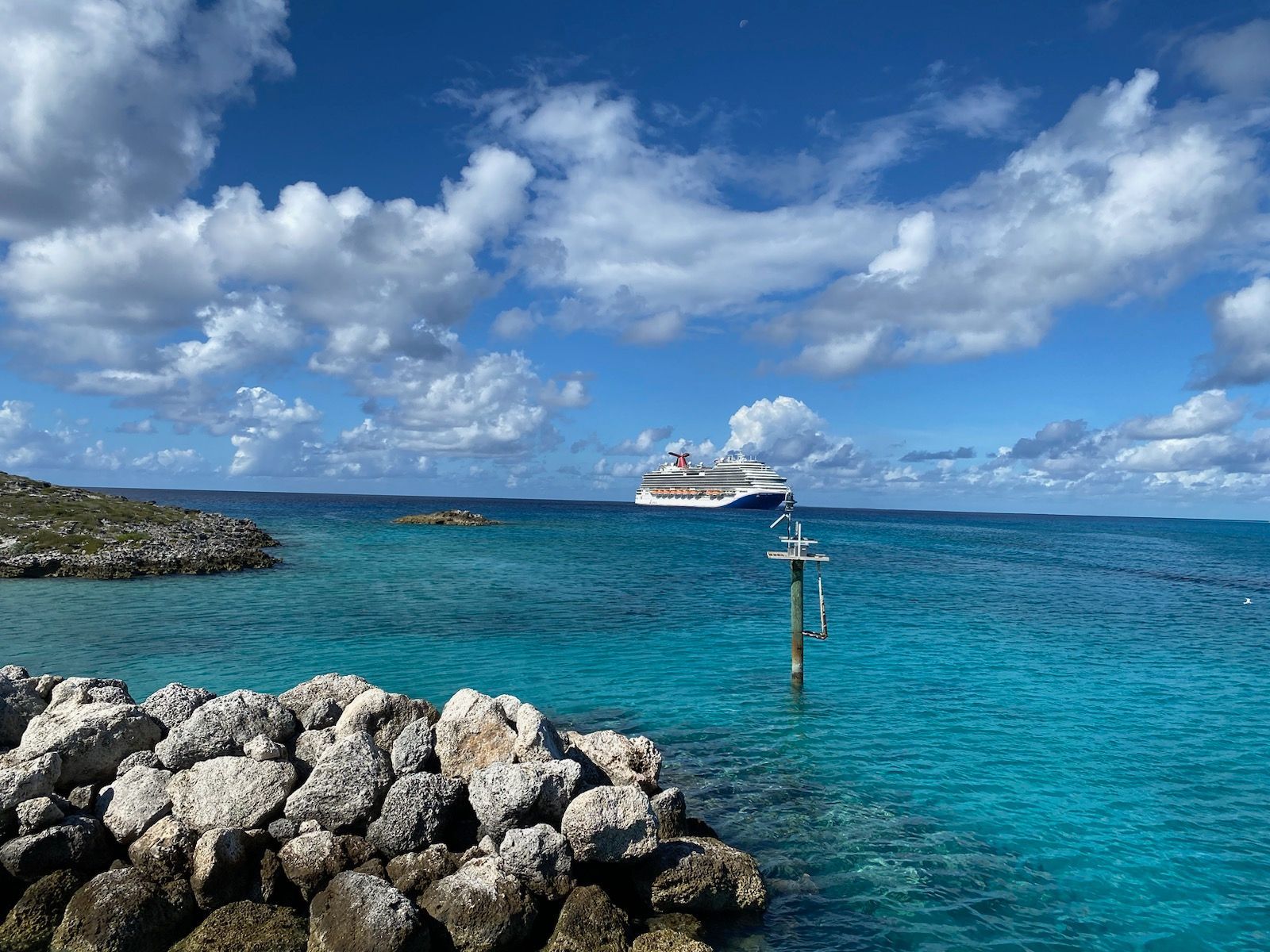 Cruise ship on turquoise water, rocky shore in foreground, blue sky with clouds.