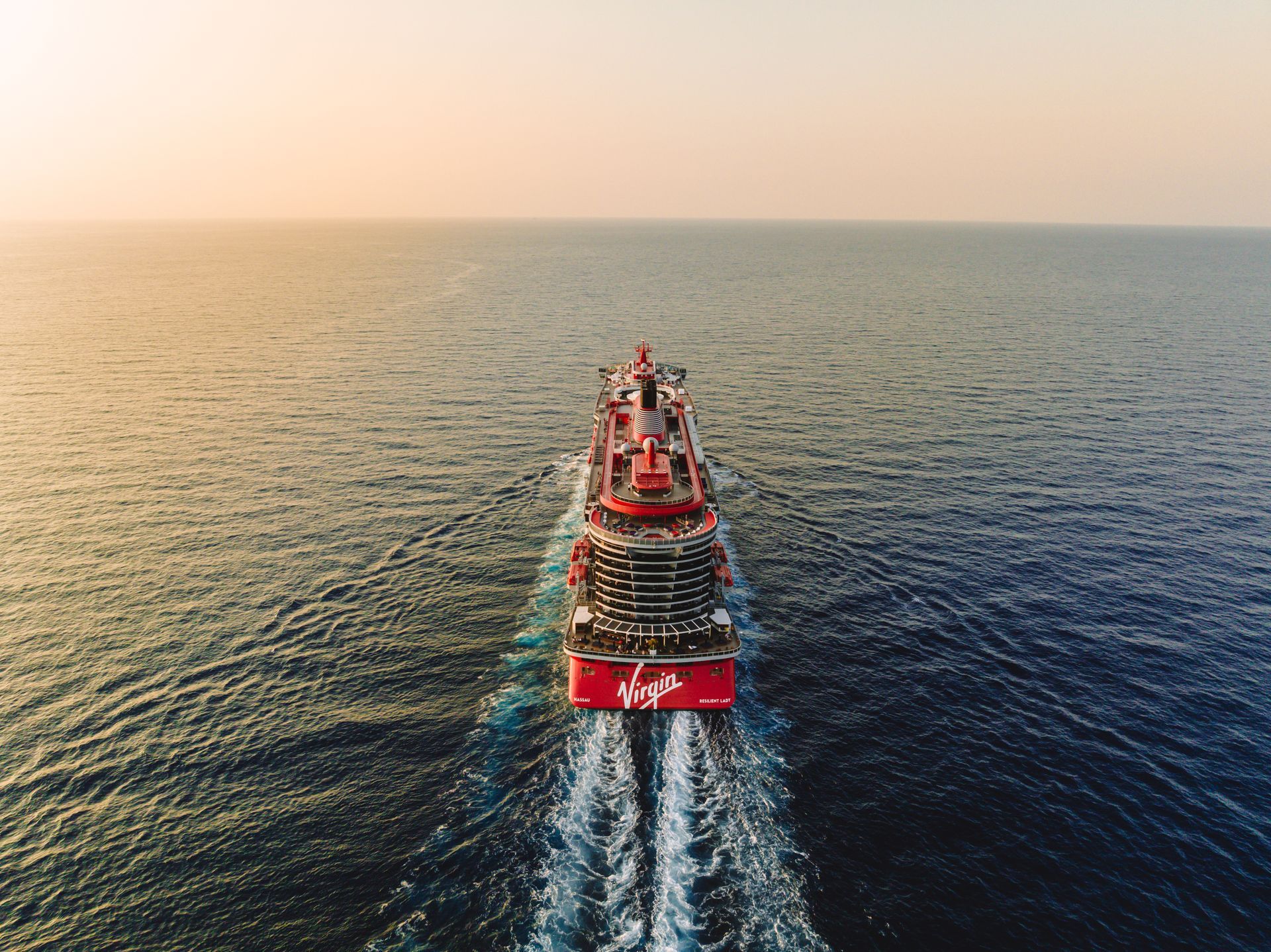 Red Virgin Voyages cruise ship sailing on the ocean, with wake, against a golden sunset.