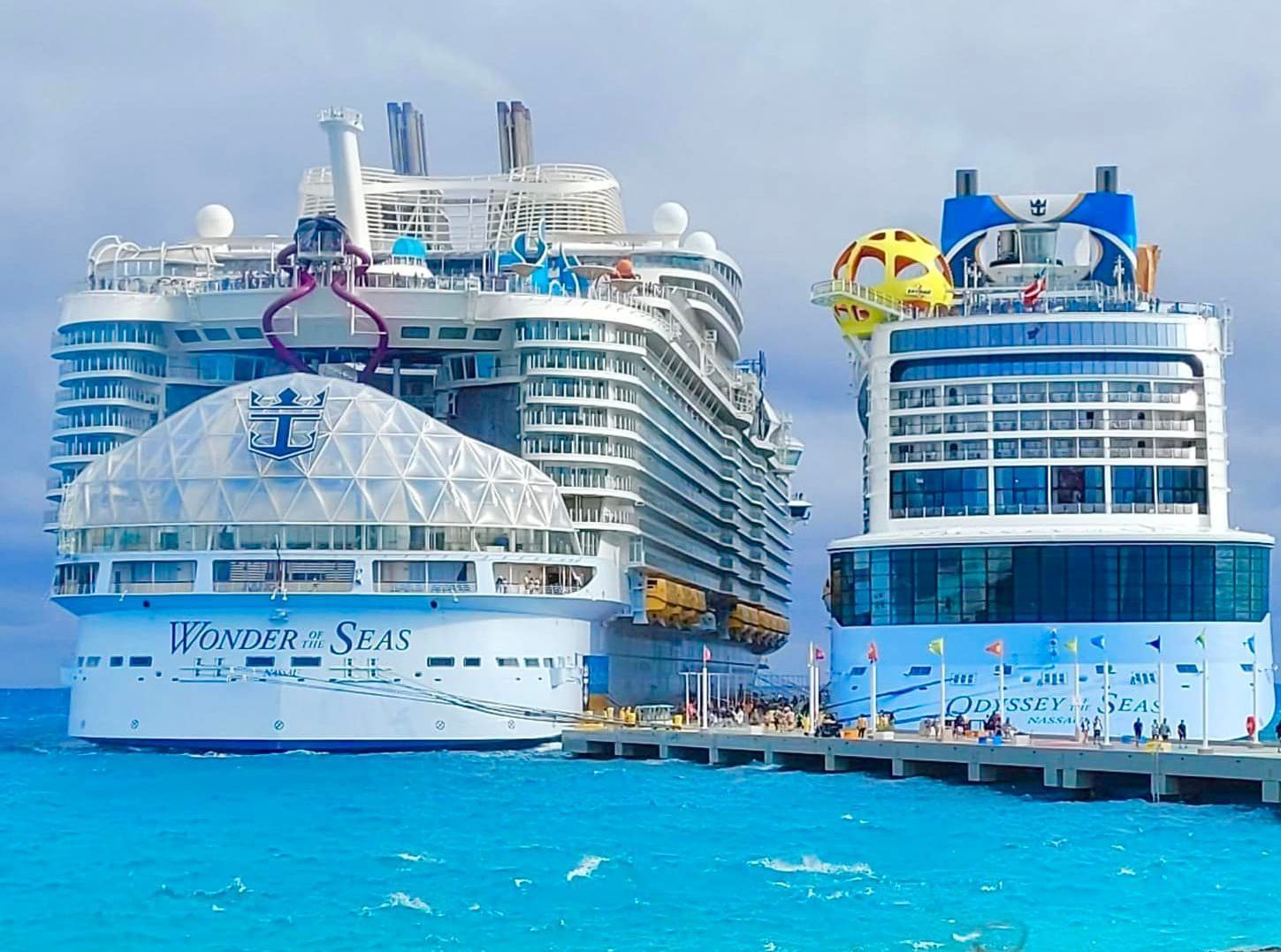 Two cruise ships docked at a pier, blue water and sky. One ship is white, the other blue with a yellow sphere.