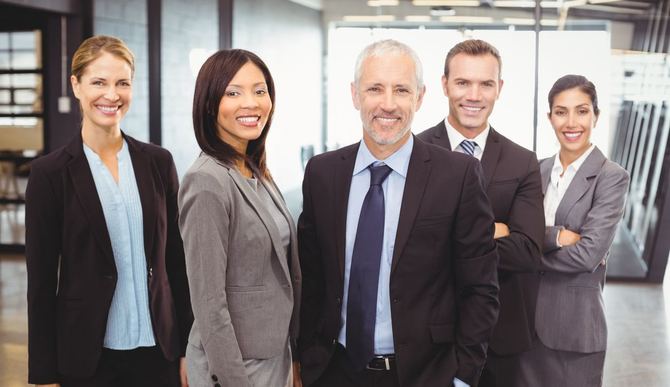 Five professionals in business attire standing and smiling in a modern office lobby