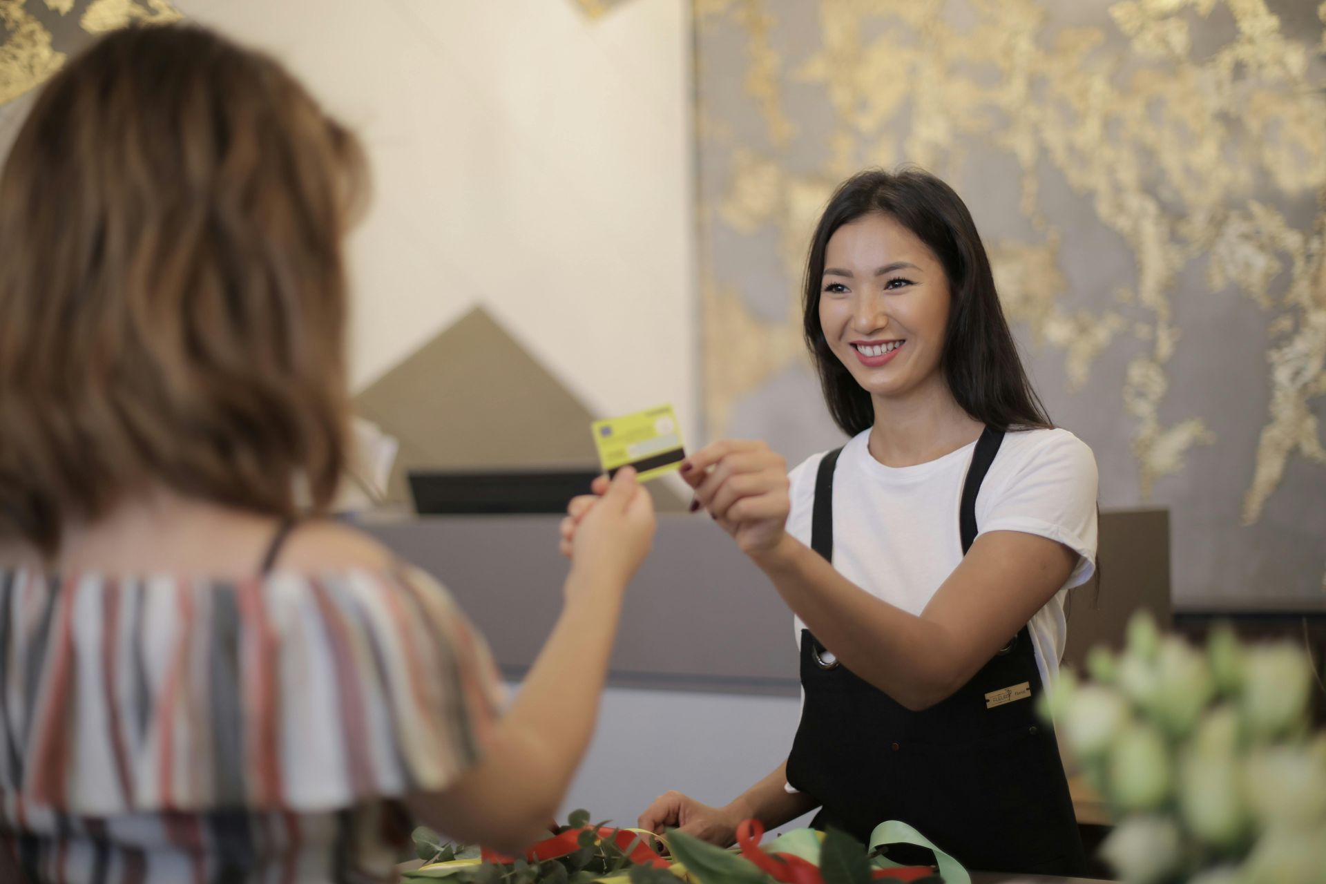 Smiling cashier hands a card to a customer at a shop counter with flowers in front.