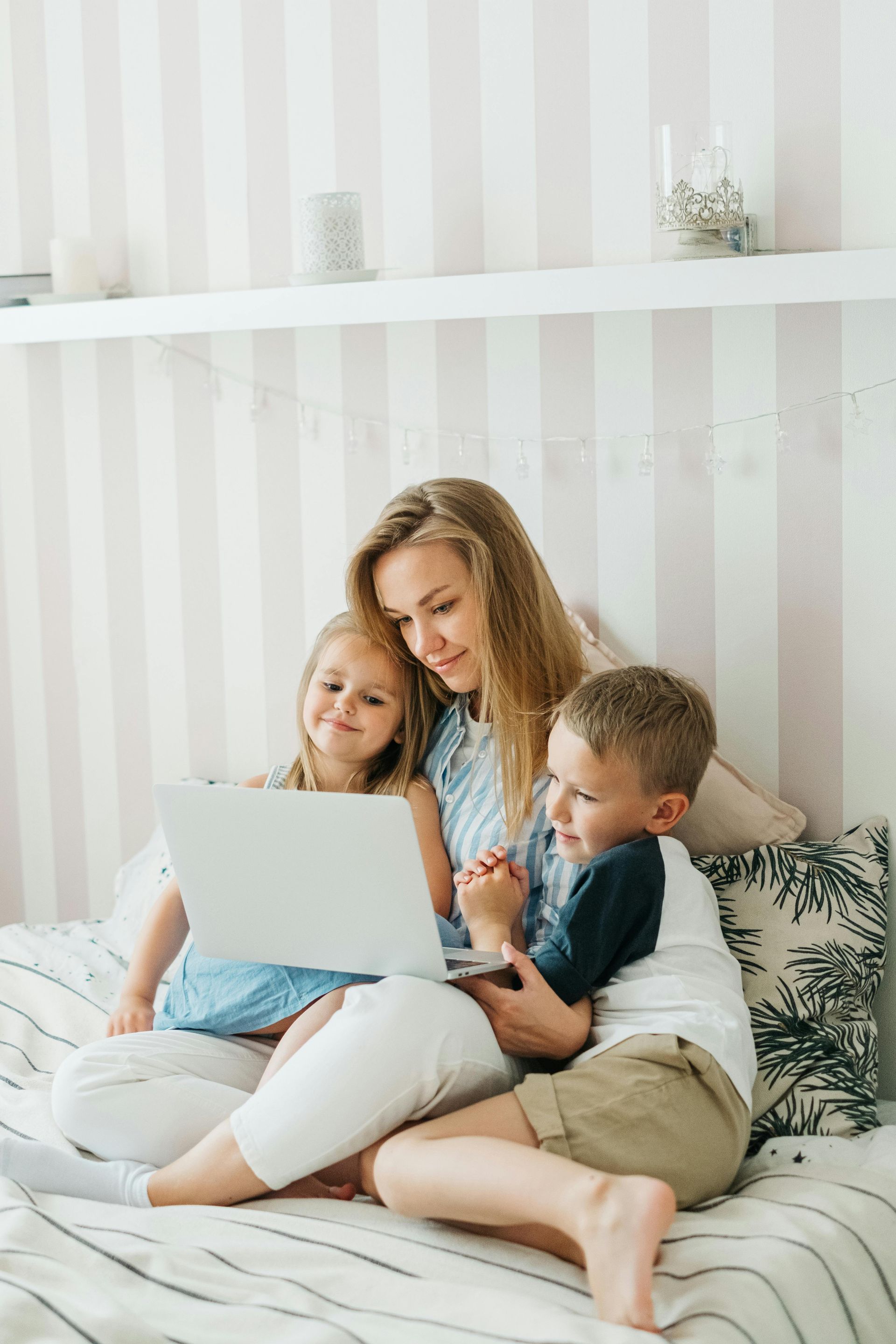 Woman and two children using a laptop while sitting on a bed in a bright bedroom