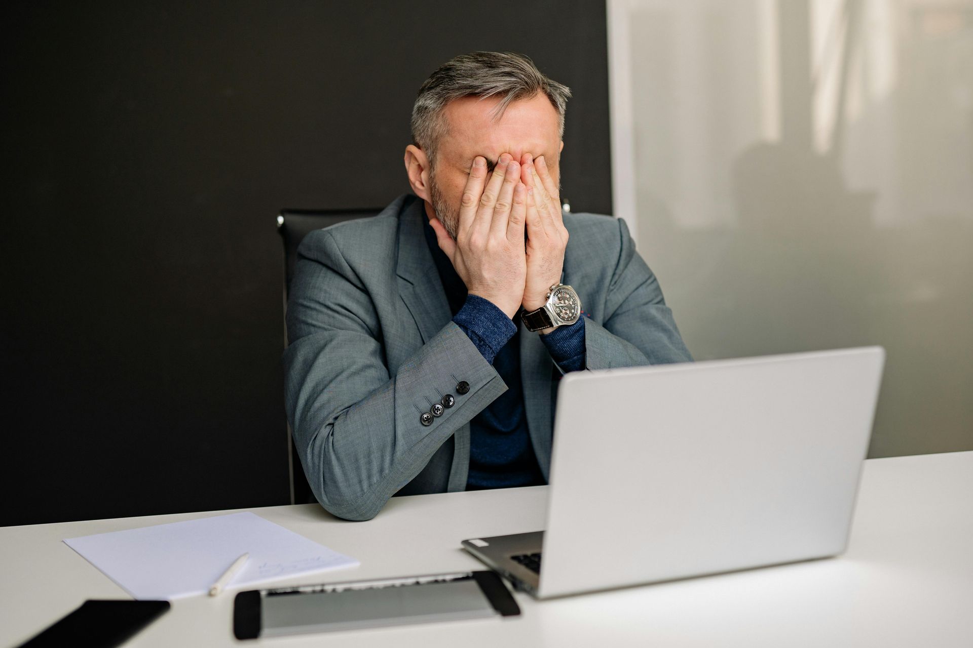 Man in a gray blazer sits at a desk, covering his face in frustration beside an open laptop.