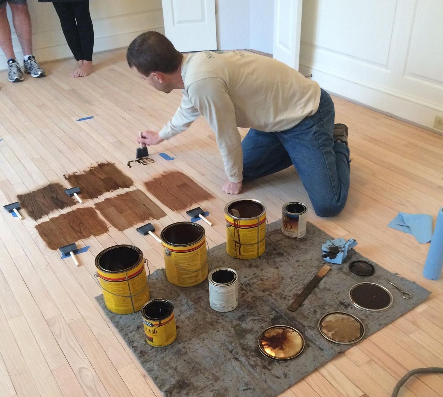 A man is kneeling on the floor surrounded by cans of paint Galebach’s Floor Finishing