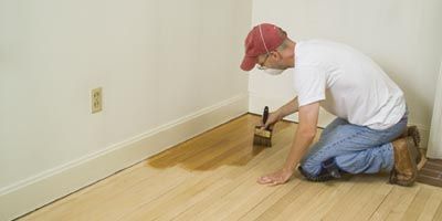 A man is kneeling down and painting a wooden floor with a brush. Galebach’s Floor Finishing