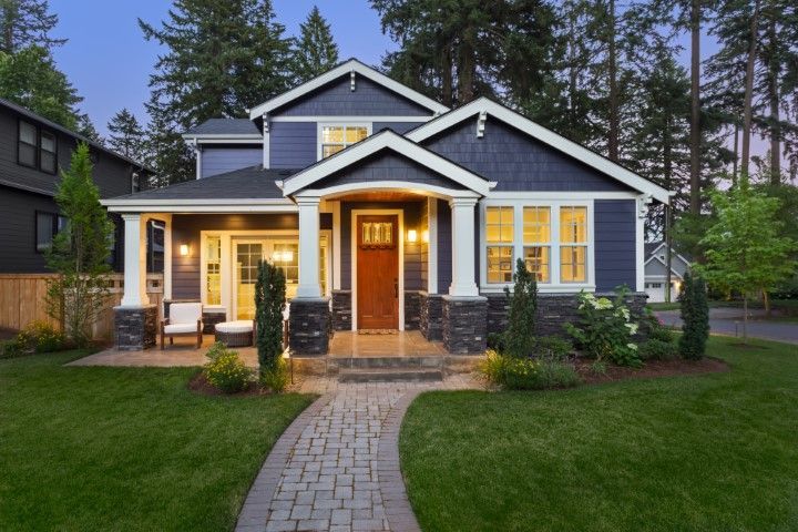 A blue, two-story craftsman-style house with a stone foundation, a wooden front door, and a brick walkway at dusk.