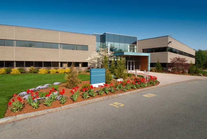 A two-story beige office building with a central glass entryway, landscaped flower beds, and a paved parking area.
