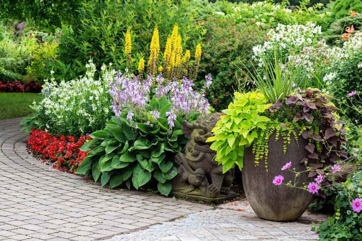 A curved stone path beside a vibrant garden bed with yellow and purple flowers, a stone statue, and a large potted plant.