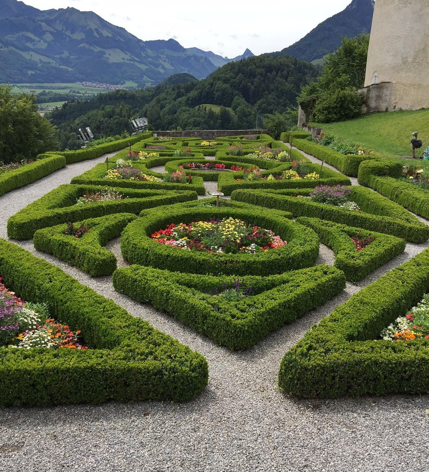 A manicured geometric garden with boxwood hedges and colorful flowers, set against a backdrop of green mountains.