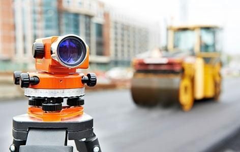 Instrumento de medición de color naranja sobre un trípode, con un rodillo compactador al fondo en un sitio de construcción.