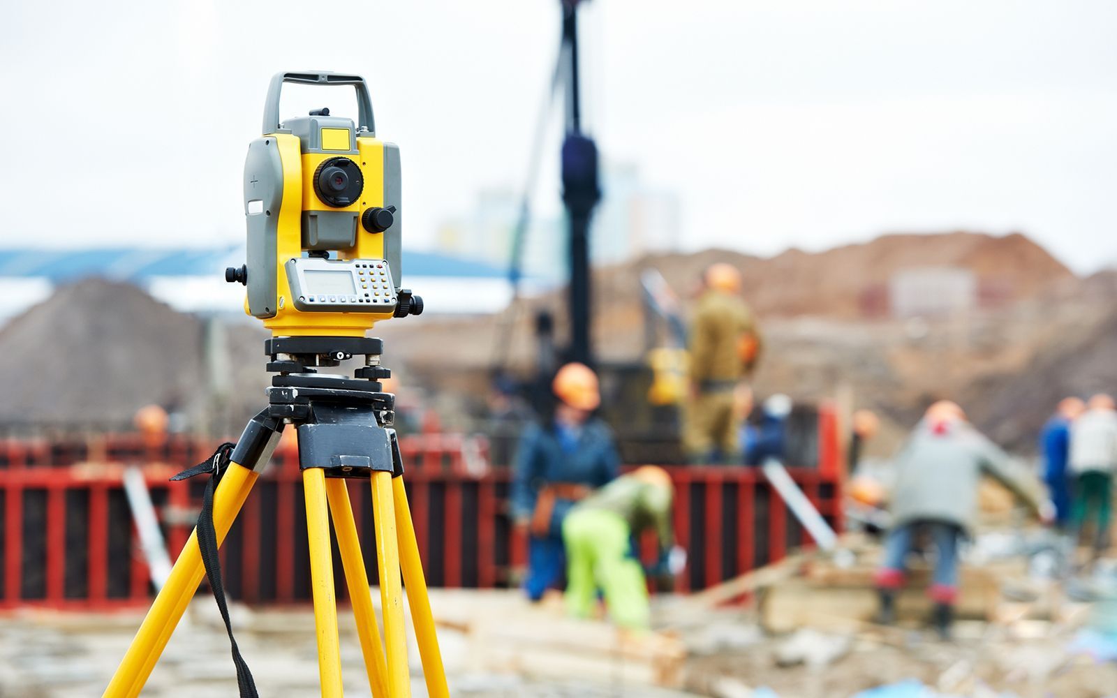 Teodolito amarillo sobre trípode en una obra en construcción; trabajadores al fondo.