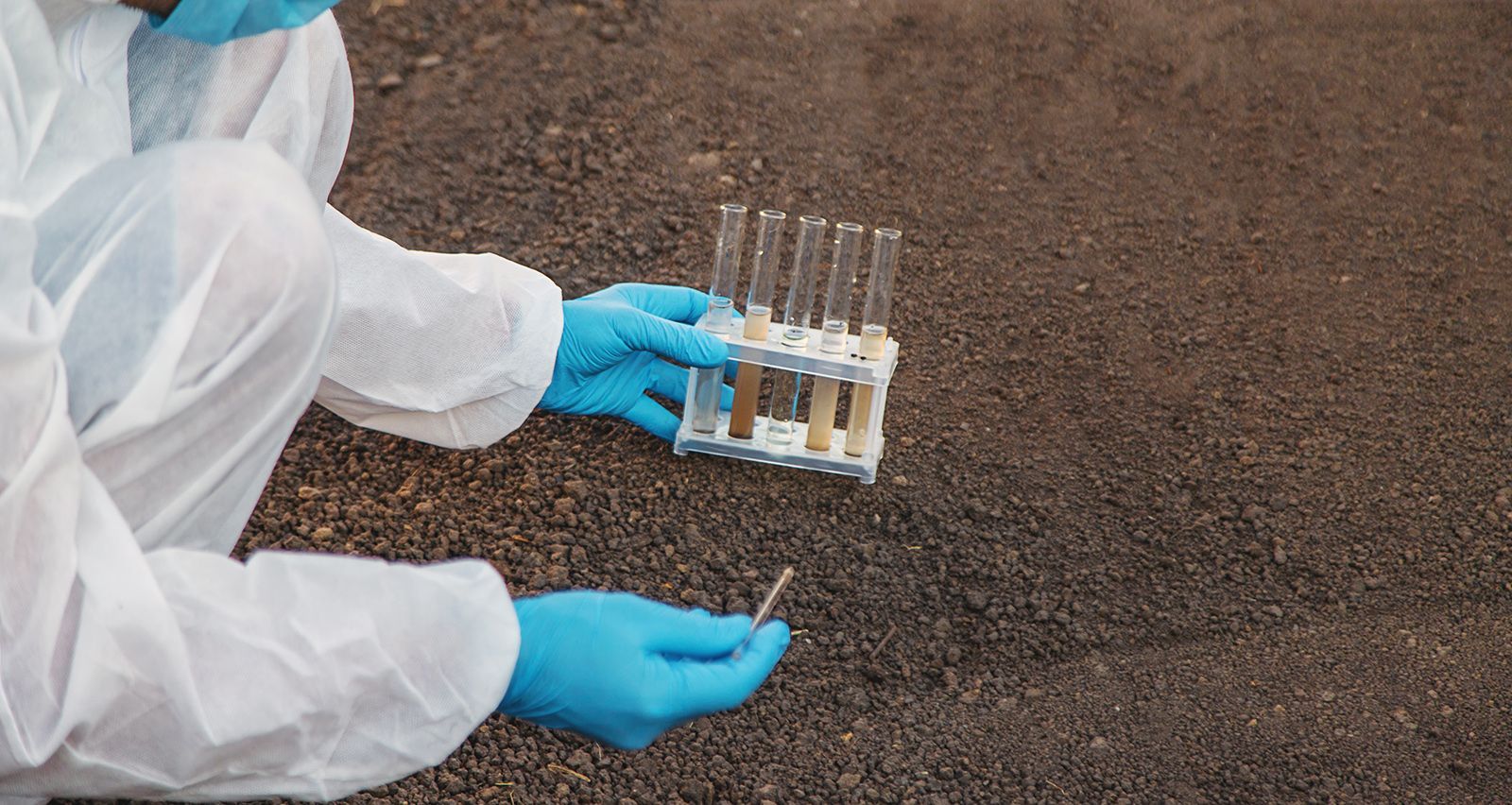 Científico con traje de protección y guantes examinando muestras de suelo en tubos de ensayo en el suelo.