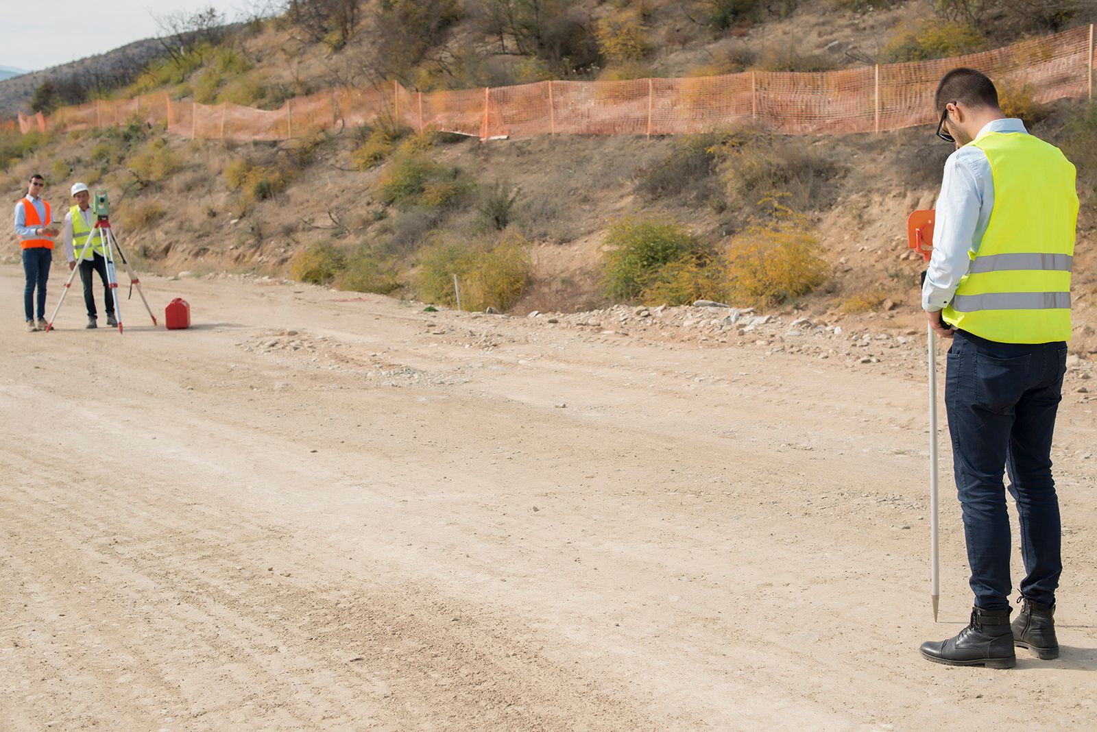 Agrimensores en un camino de tierra, uno sosteniendo una vara de medir, otros operando equipo de topografía; cerca naranja al fondo.
