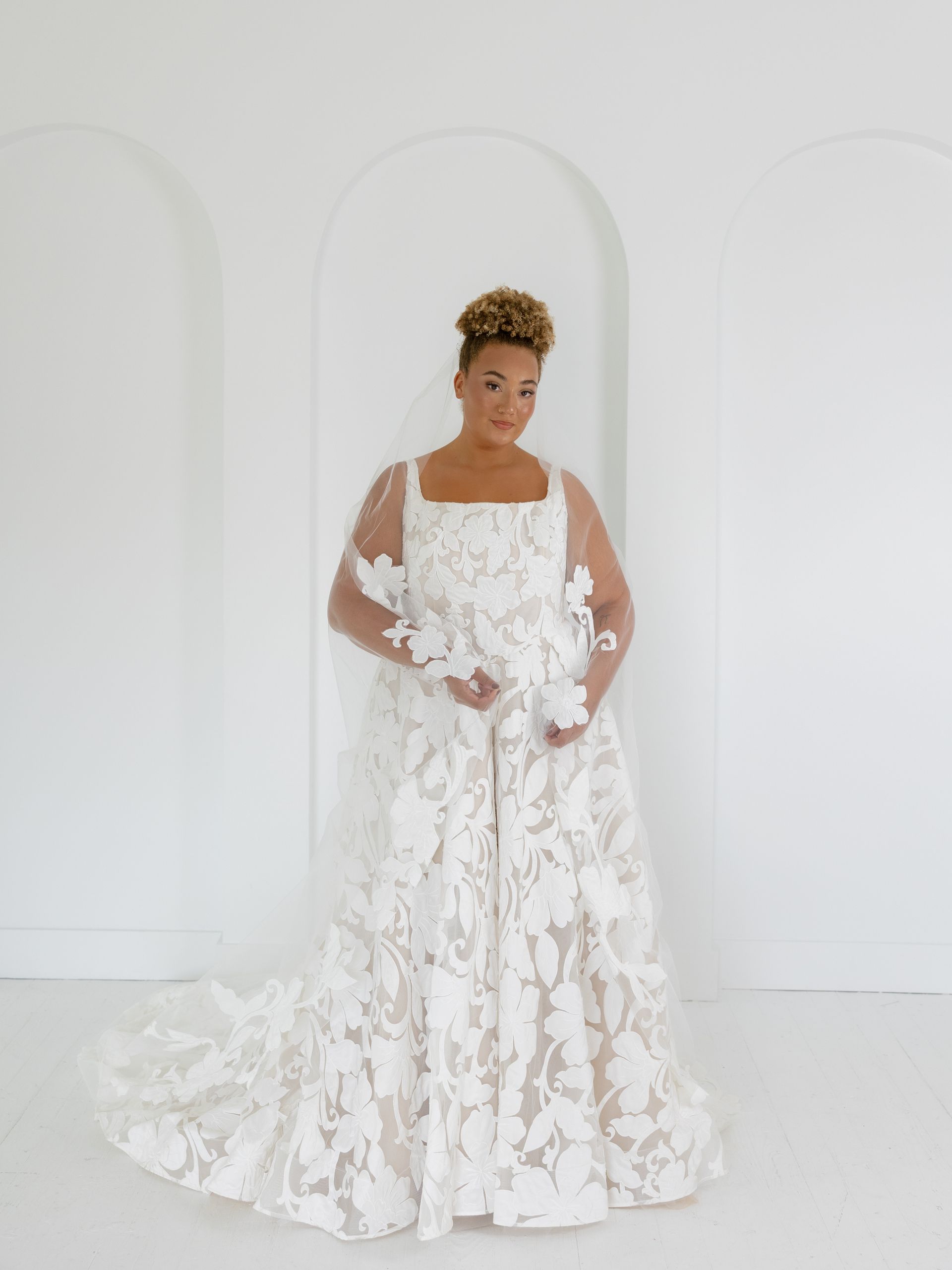 Woman in floral wedding dress and veil stands in a white arched room.
