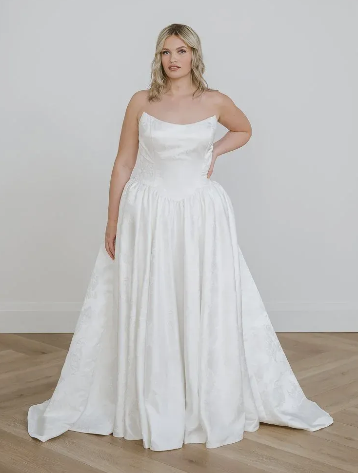 Bride in white wedding dress, holding up skirt, standing in a studio.