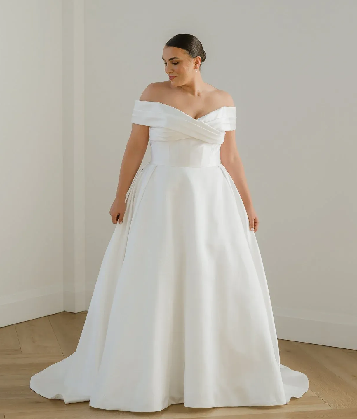 Woman in wedding dress, standing indoors with hands on hips. Dress is white with lace detailing, and she is looking away.