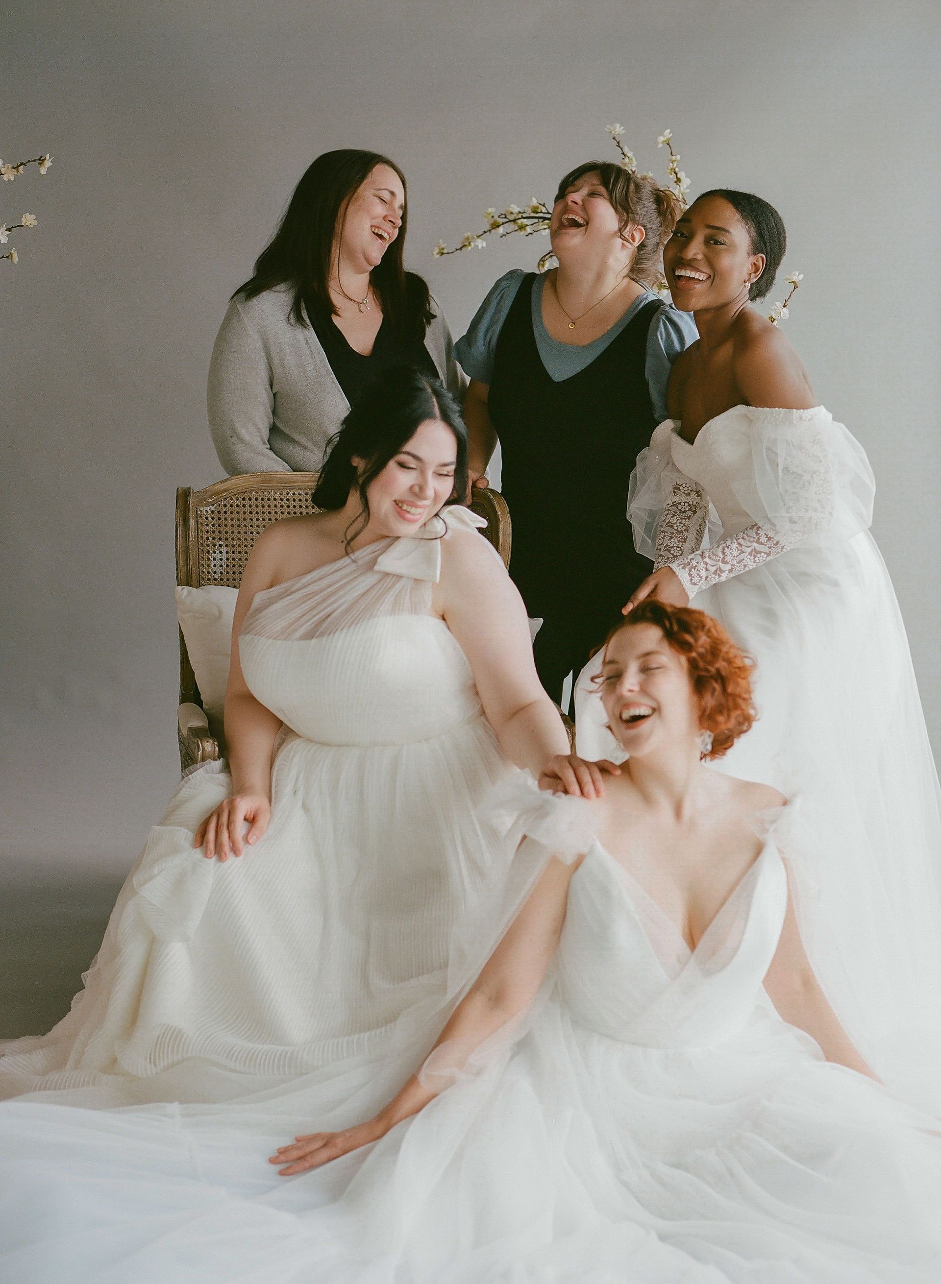 Six women in wedding dresses laugh together. Gray background, floral accents.