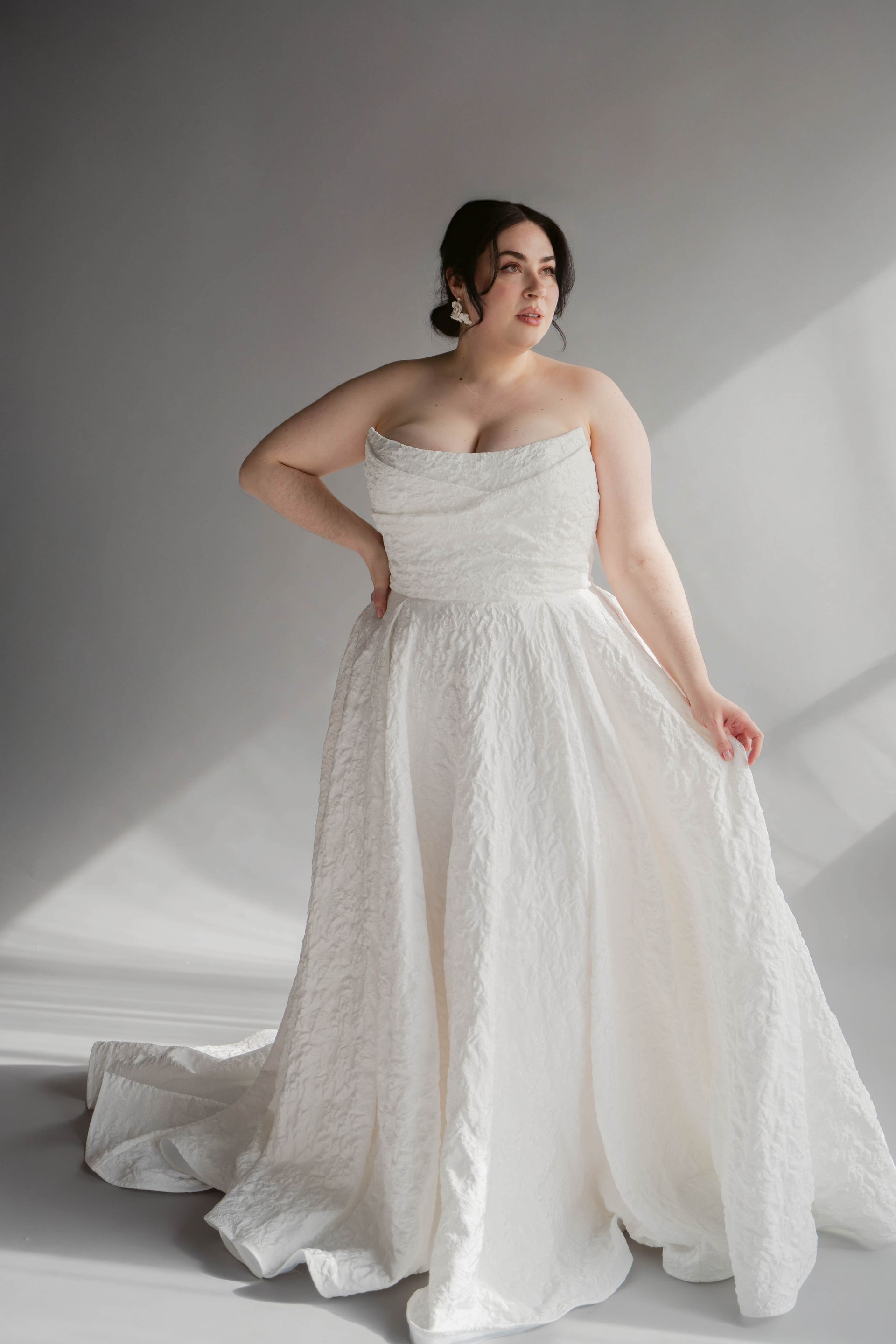 Woman in a white wedding gown with a full skirt and train, posing with hand on hip.
