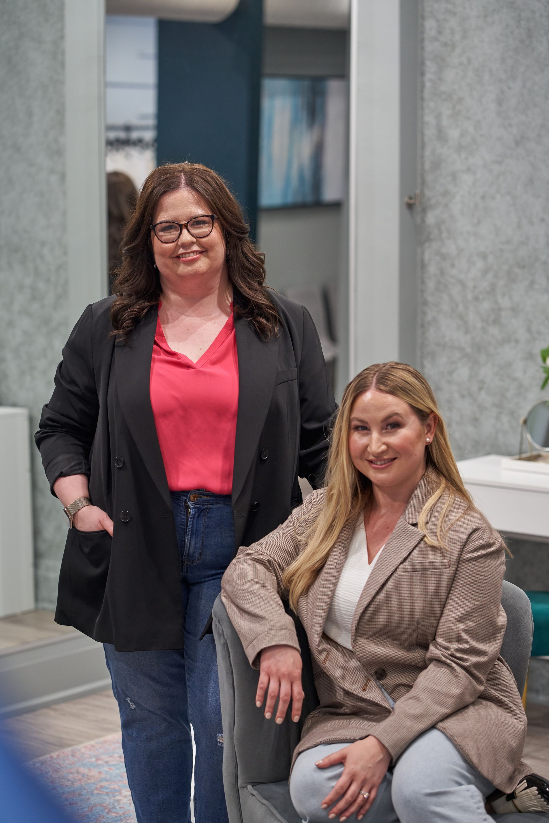 Two people wearing white off-the-shoulder wedding dresses pose against a gray backdrop.